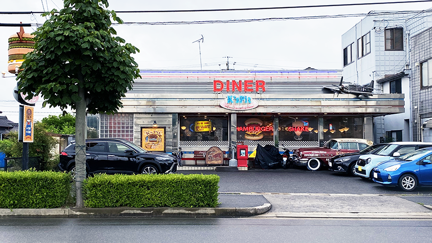 ケーズピットの店舗外観。レトロなアメリカンダイナー風の建物で、軒先に飾られている外車や看板上の飛行機にこだわりを感じられる width=