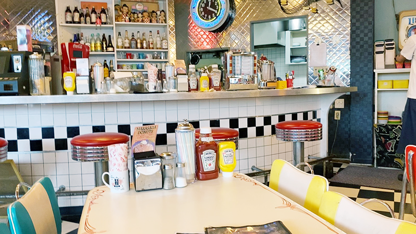 アメリカンダイナーを彷彿とするカウンター。テーブルにはケチャップやマスタードの調味料が所狭しと並び、壁にはネオンが輝くインテリアやチェック模様の銀の壁がある