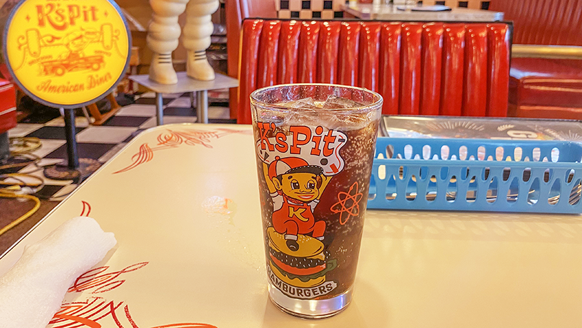 食前に運ばれてきたコーラの写真。容器にはケーズピットオリジナルキャラクター(赤色のつなぎと帽子を身につけたおじさん)が描かれている